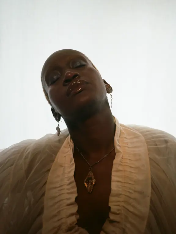 Portrait of person with closely cropped hair, eyes closed, wearing translucent ruffled garment and pendant necklace, head tilted back, soft lighting from above, minimalist pale background creating calm, atmospheric composition.