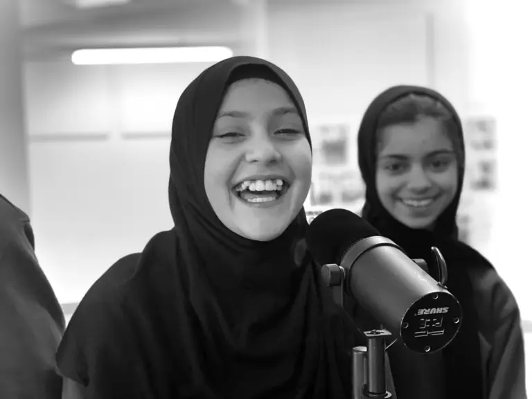 a young woman with a headscarf smiling into a microphone, with another young girl smiling behind her. Black and white