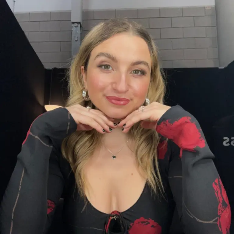 Chloe resting her chin on both of her hands and giving a slight smile. She is wearing a black top with red roses on it, she has blonde hair.