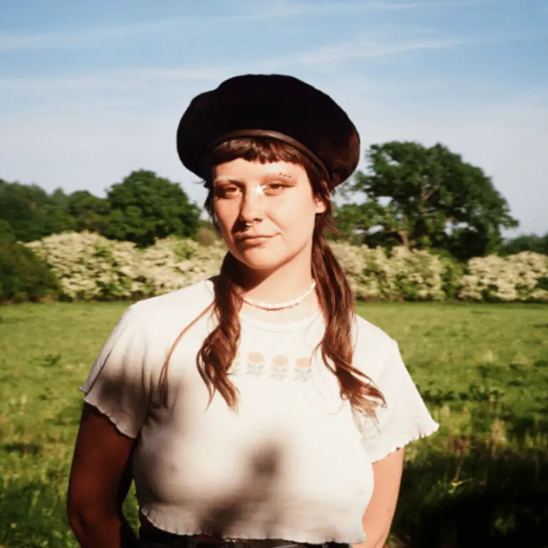 Mollie wearing a white top and a hat in a sunny field, with trees and grass and bushes in blossom behind her