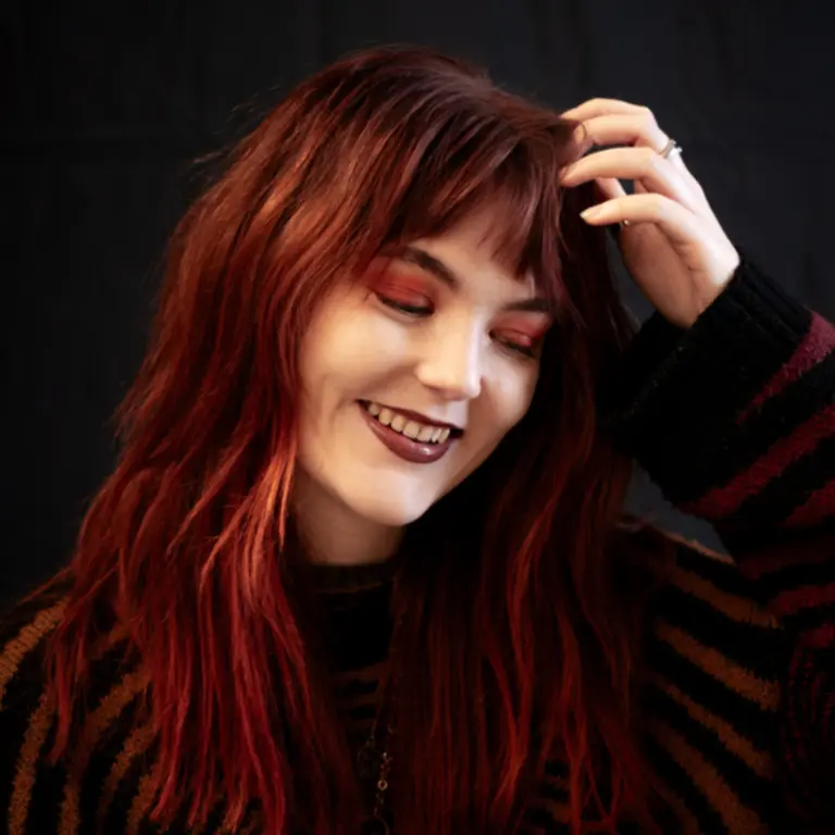 Jess smiling with her hand running through her hair with her eyes closed, looking to the bottom right of the camera. Her hair is auburn/red and matching her red top with black stripes.