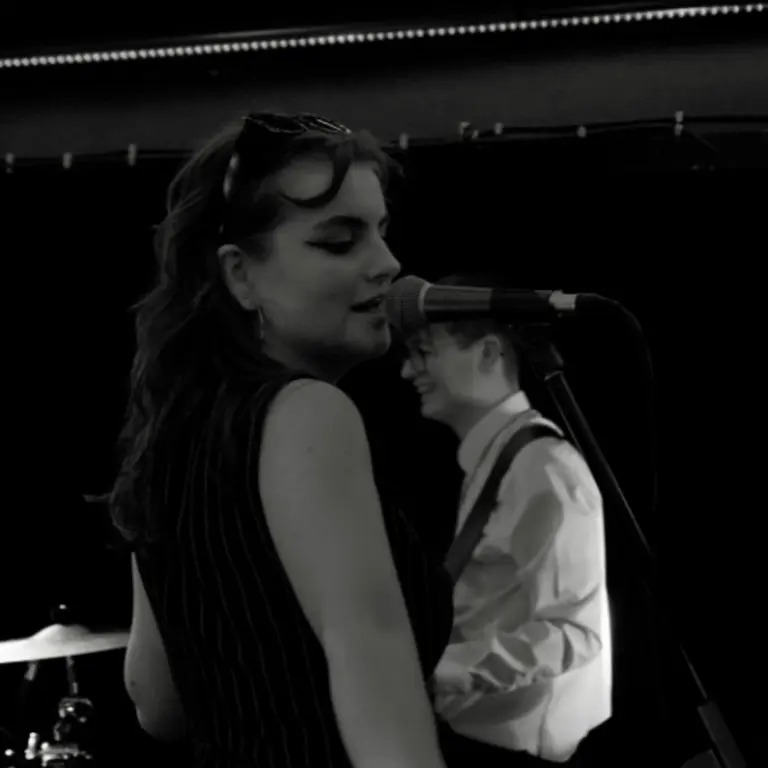 Isabel singing into a microphone from the side, wearing a sleeveless top and glasses on her head holding her hair back. 