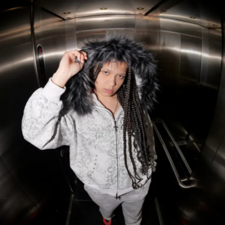 JayaHadADream stands in a lift, wearing a light patterned tracksuit with a furry hood pulled up. They look directly at the camera with a serious expression.