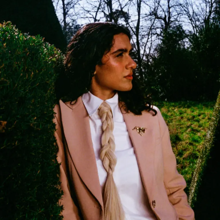 Girl in dustry pink jacket, white shirt, ponytail as a tie, leans against a hedge and looks pensively to the right of screen