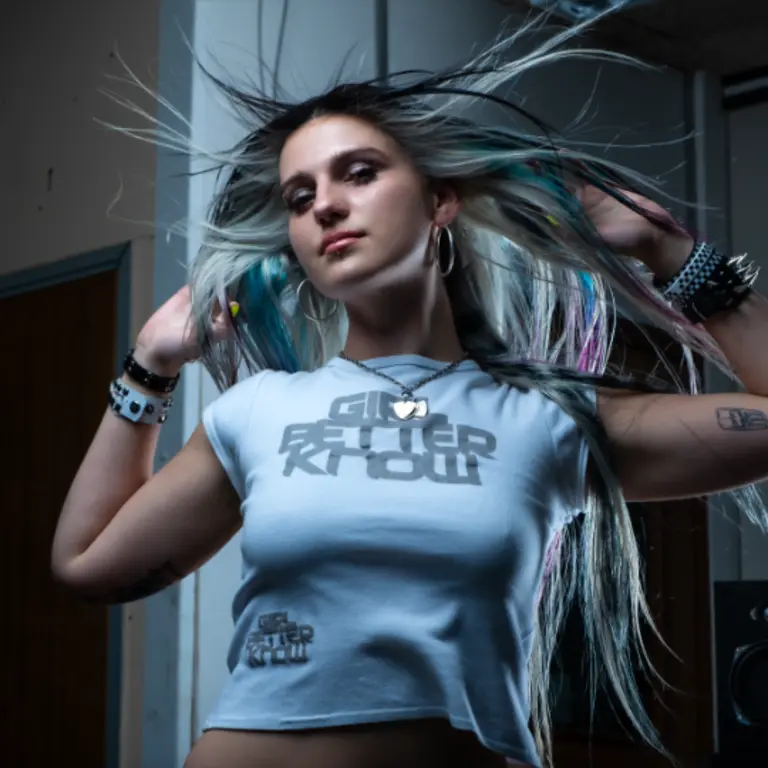 coupdekat with her hair flicked up by her hands in a dynamic manner, looking down at the camera, in an industrial background wearing a white top
