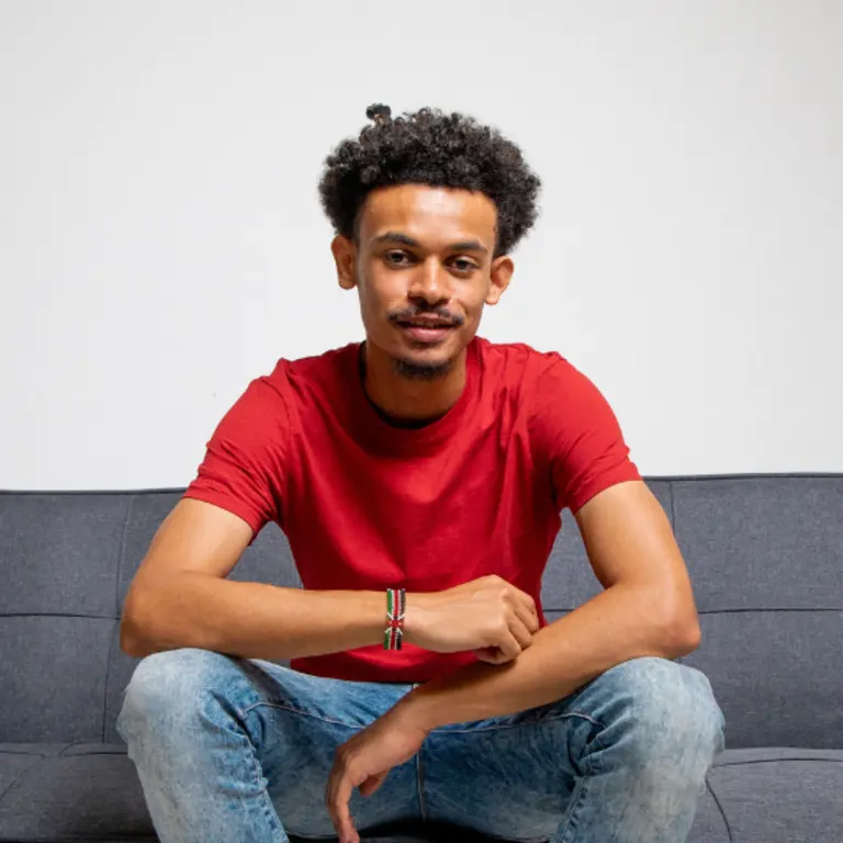 Mwathi smiling sat on a sofa, with a red top and blue jeans, resting his elbows on both of his knees. 