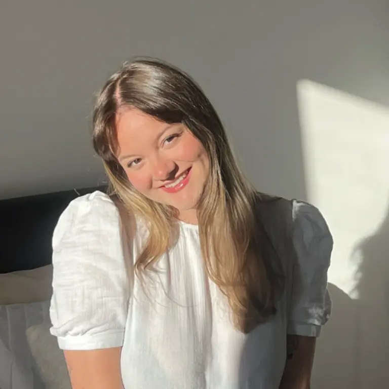 Isobel smiling in a sunny room, blonde hair and a white top, with a square of sunlight from the window as a shadow behind her