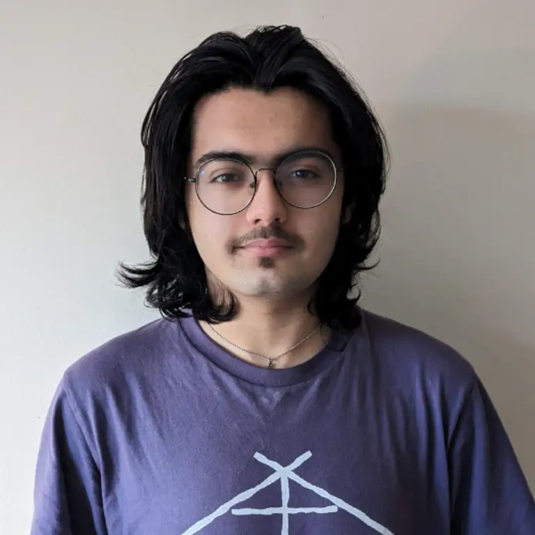 Tom Rabbani headshot, purple t-shirt on in front of a wall, neutral expression