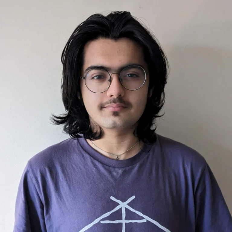 Tom Rabbani headshot, purple t-shirt on in front of a wall, neutral expression