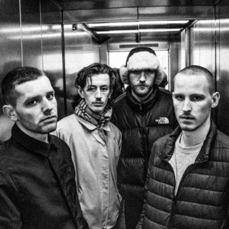 All 4 members looking expressionless at the camera wearing jackets and coats, black and white