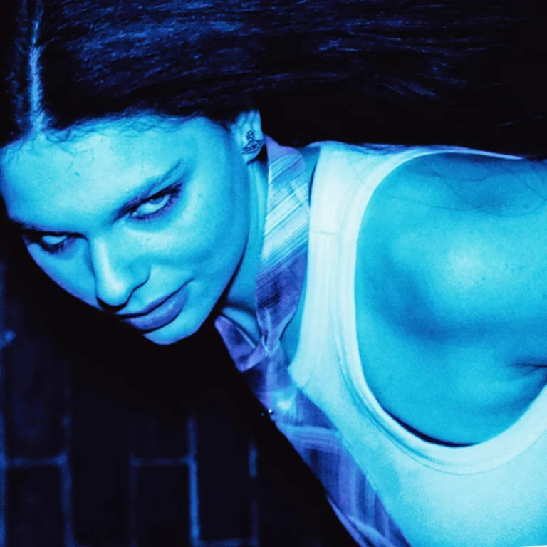 Boudicca with long dark hair, wearing a sleeveless white top, leans forward and looks intently at the camera. The image is heavily tinted in blue, creating a dramatic, high-contrast effect.