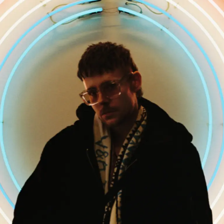 Person with a jacket and scarf inside a thin circle of blue/green light, wearing sunglasses, looking at the camera