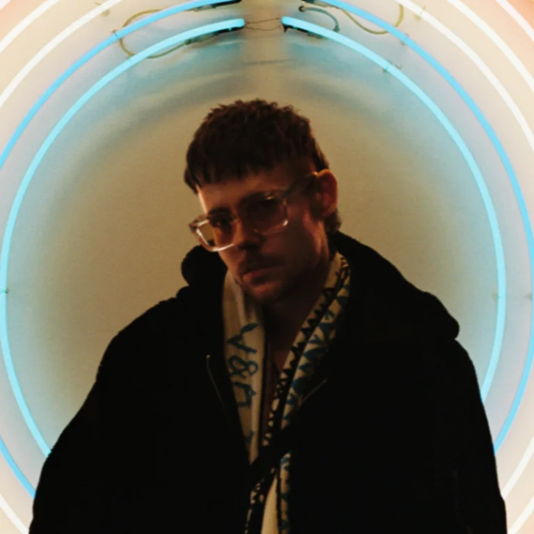 Person with a jacket and scarf inside a thin circle of blue/green light, wearing sunglasses, looking at the camera