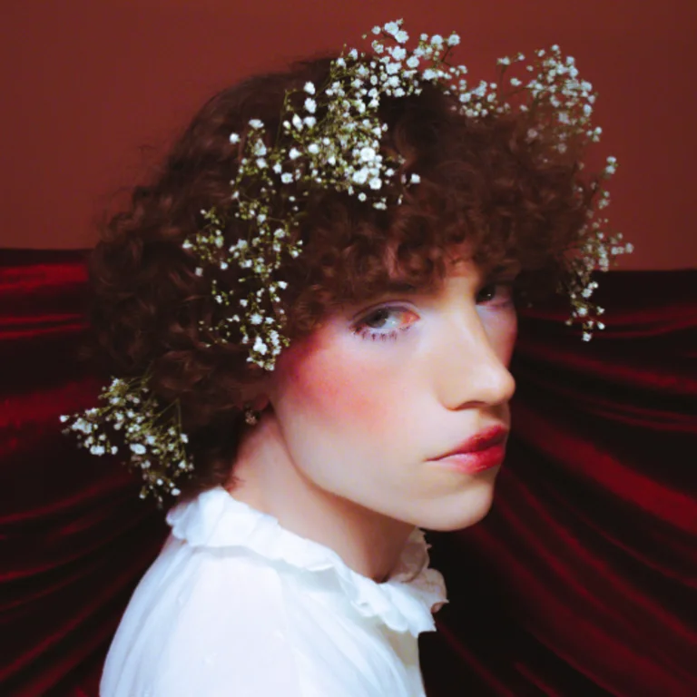 Garret wearing a crown and blusher, looking over his shoulder at the camera, with a red velvet background and a white shirt
