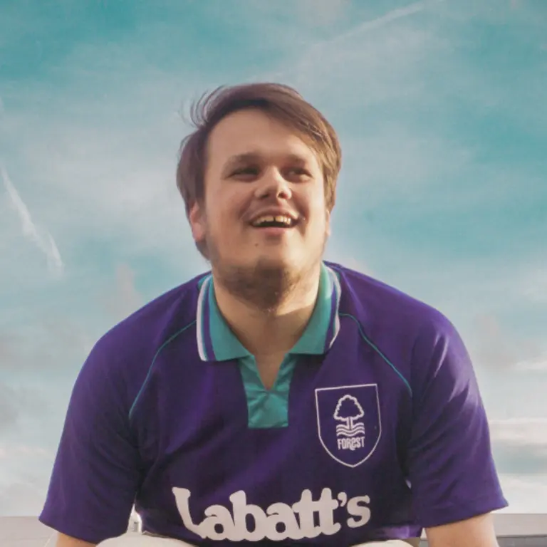 An image taken from below, with olly smiling but looking over the camera with a blue sky with wispy white clouds. He is wearing Nottingham Forest FC's third kit, in purple with a turqoise colour