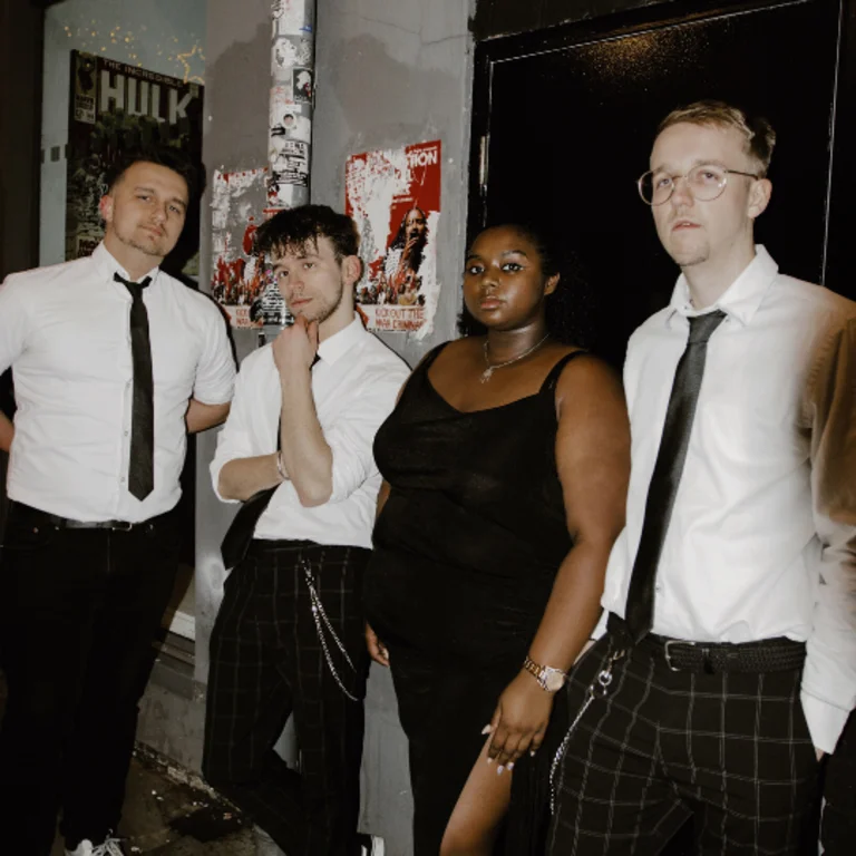 all 4 members stood around the camera in a semi circle. All the boys in smart trousers with white shirts and ties, the female singer wearing a black dress