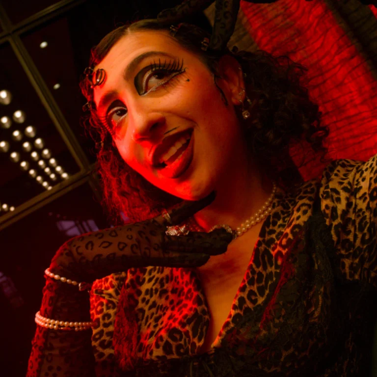 Rivkala poses under red lighting, smiling with her mouth slightly open. She wears dramatic eye makeup, a leopard-print outfit, pearl jewellery, and sheer patterned gloves, holding one hand playfully to her chin.
