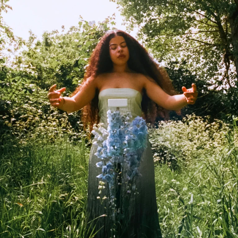 Afromerm with long curly hair wearing a strapless floral dress, standing outdoors in a lush garden setting with arms outstretched, surrounded by trees and wildflowers.