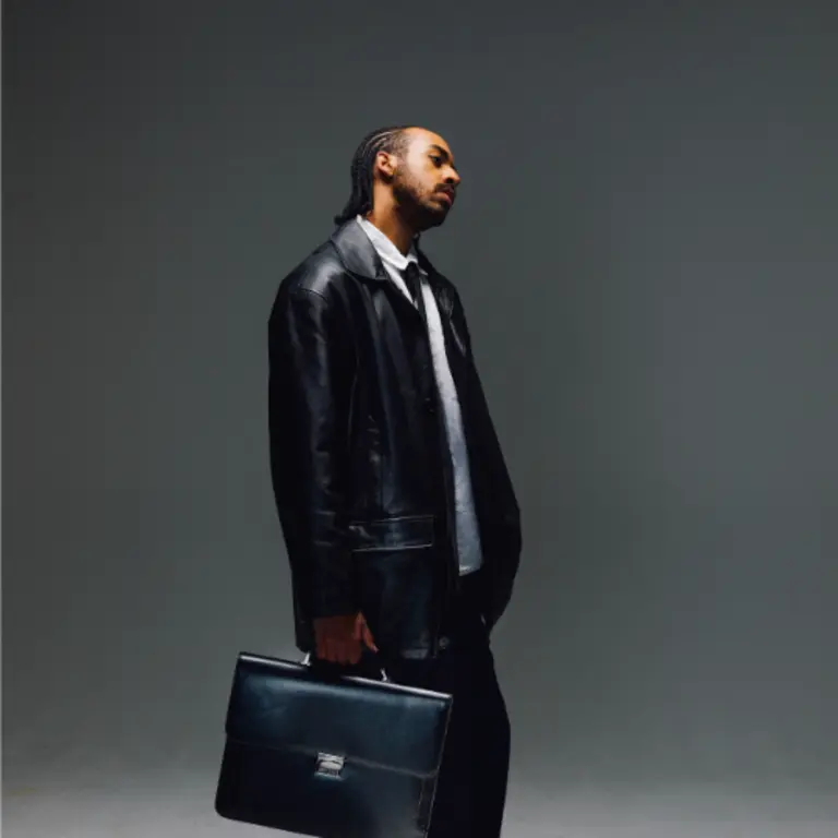 simon a holding a briefcase wearing a black leather jacket and a white shirt, facing to the right