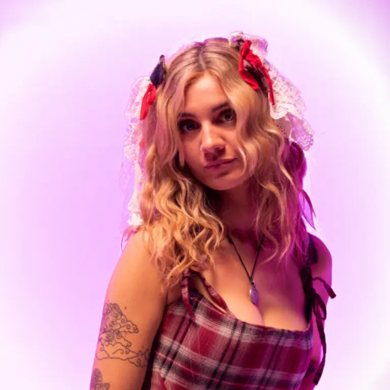 Mali Haf in a red tartan dress and long blonde hair over her shoulders, looking over her right shoulder at the camera, with a purple effect around her