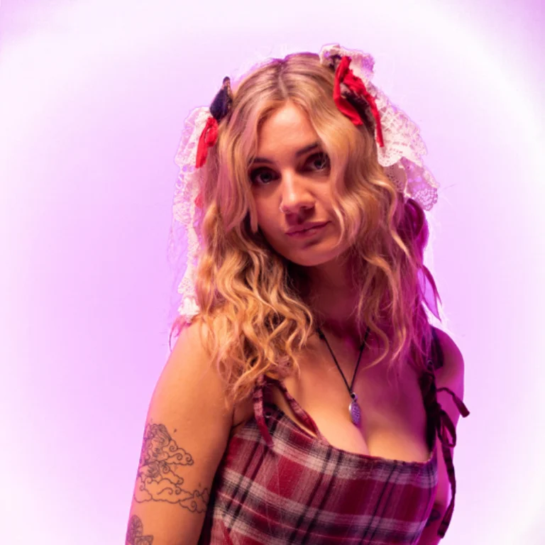 Mali Haf in a red tartan dress and long blonde hair over her shoulders, looking over her right shoulder at the camera, with a purple effect around her