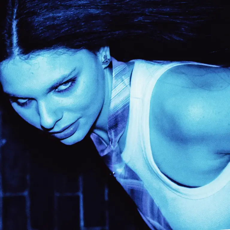A woman with long hair poses in intense blue lighting, wearing a sleeveless top. Her expression is mysterious and confident, set against a dark background.