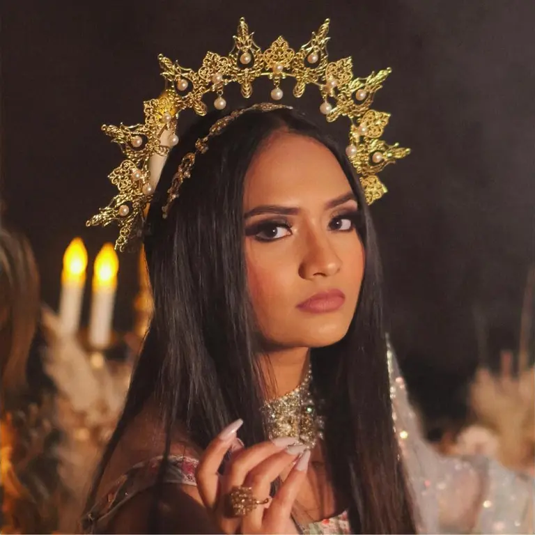 A woman with long dark hair wears an ornate gold crown and a jeweled necklace. Her expression is serious, set against a soft, dimly lit background.
