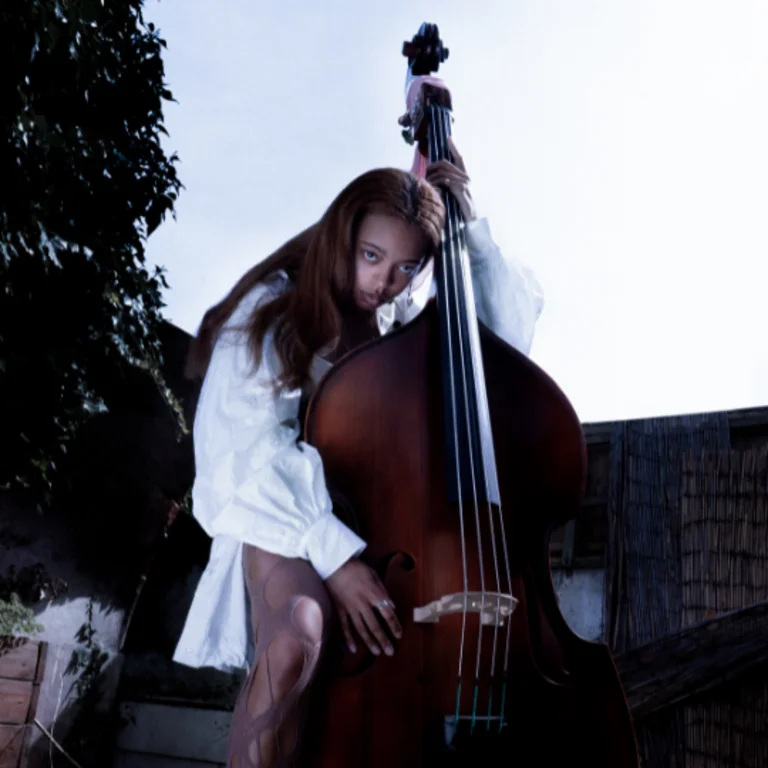 Woman with long black hair holding a cello/double bass, arm, leg and head resting on it, outside with a grey effect