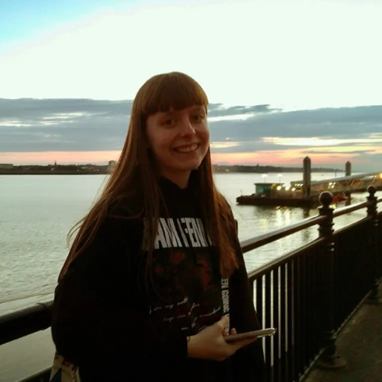 Ellen in front a pier by the sea as there is an orange sunset behind her. She is smiling and wearing a hoodie