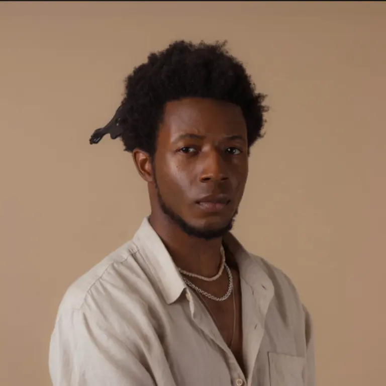 Wheelan wearing a cream shirt with two necklaces, staring down the camera in a thoughtful way. He has a sand coloured professional backdrop