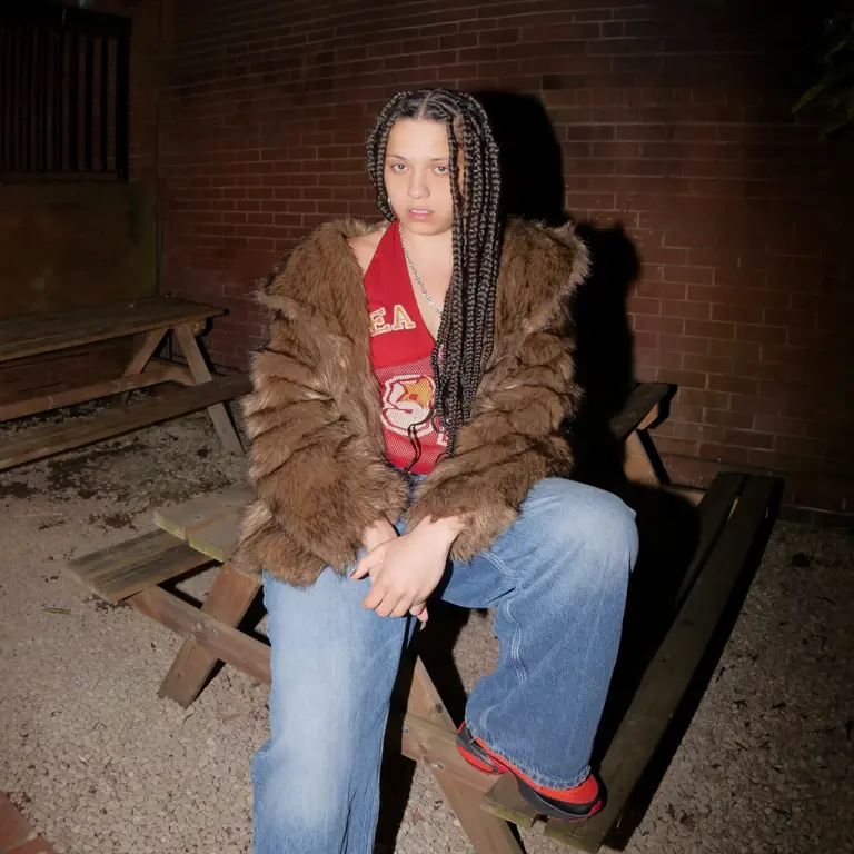 A young woman with braided hair sits on a wooden picnic table outdoors at night. They wear a fur coat, red patterned top, and jeans, exuding a relaxed vibe.