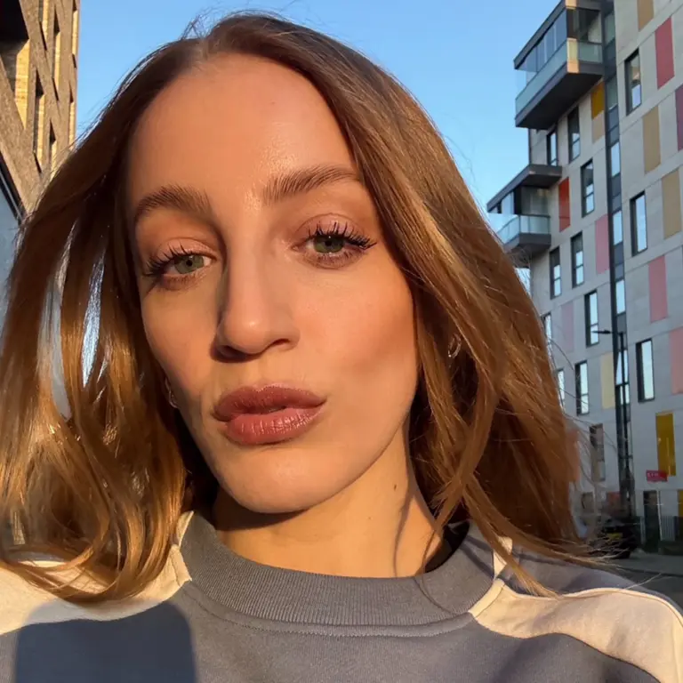 Joss has light brown hair and takes a selfie outdoors at golden hour. Urban background with modern buildings, soft orange lighting, and a calm expression.