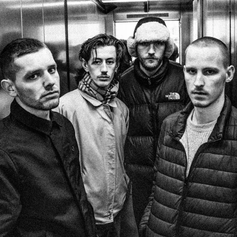 Black and white photo of four young men standing in an elevator. They wear casual winter clothing, expressing serious, contemplative expressions.