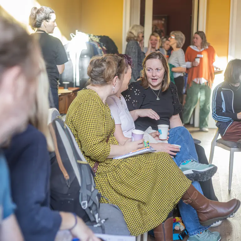 A picture from the energiser event in bristol, adults entering a hall and chatting with each other on seats or stood near the door
