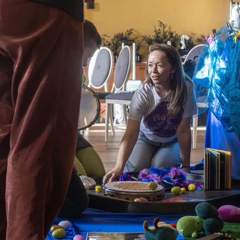 a woman on her knees smiling and talking to someone off screen, in front of them are all sorts of toys and musical instruments laid out on a blue sheet