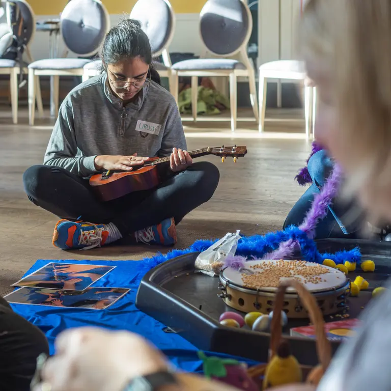 a girl sat on the floor of a hall with a ukelele, and surrounded by other musical instruments and toys