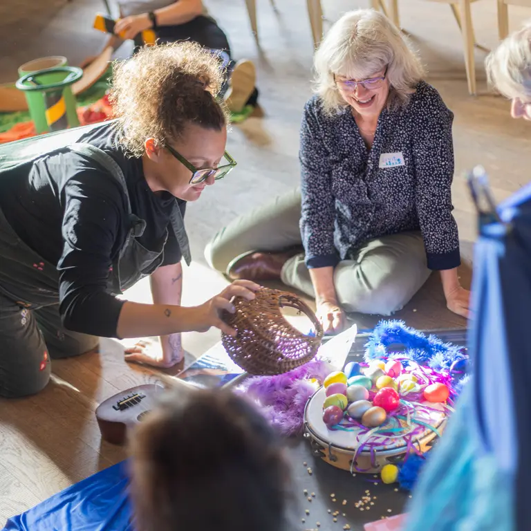 3 adults smiling and kneeling on the floor, playing with small percusive shakers and other musical toys on a blue sheet