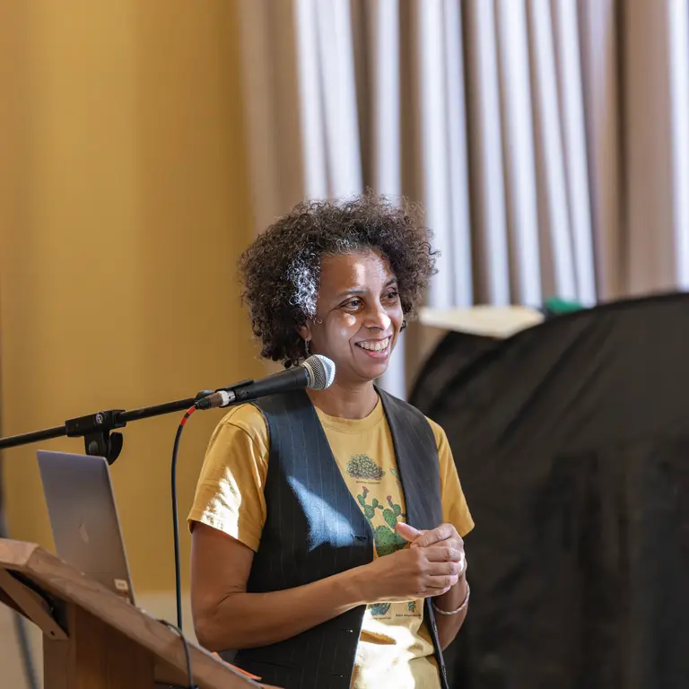 Dr Adesola Akinleye smiling in front of a microphone and lecturn, with the sun hitting her face. She is clasping her hands in front of her yellow shirt