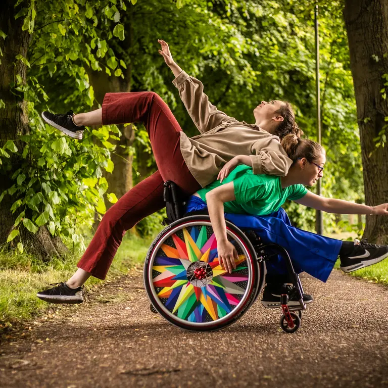 Two participants create a balanced movement shape outdoors, with one seated in a wheelchair and the other leaning back across them, arms extended, on a tree‑lined path.