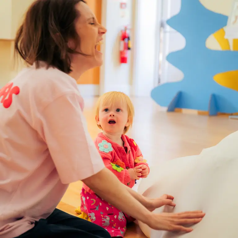 An adult woman kneeling on the floor and laughing while a baby looks up at her