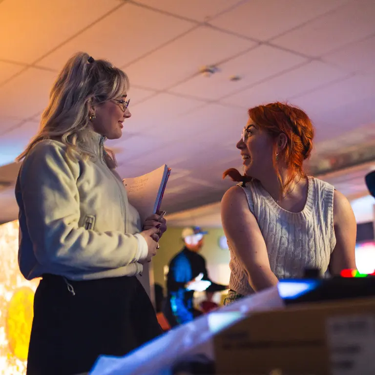 Three people gather around sound equipment in a low‑lit room, with one holding a microphone while two others talk nearby, surrounded by screens, cables and workshop materials.