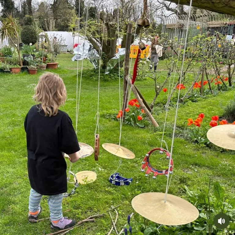 A child stnads in a garden with flowers, and cymbols hanging in front of them on strings