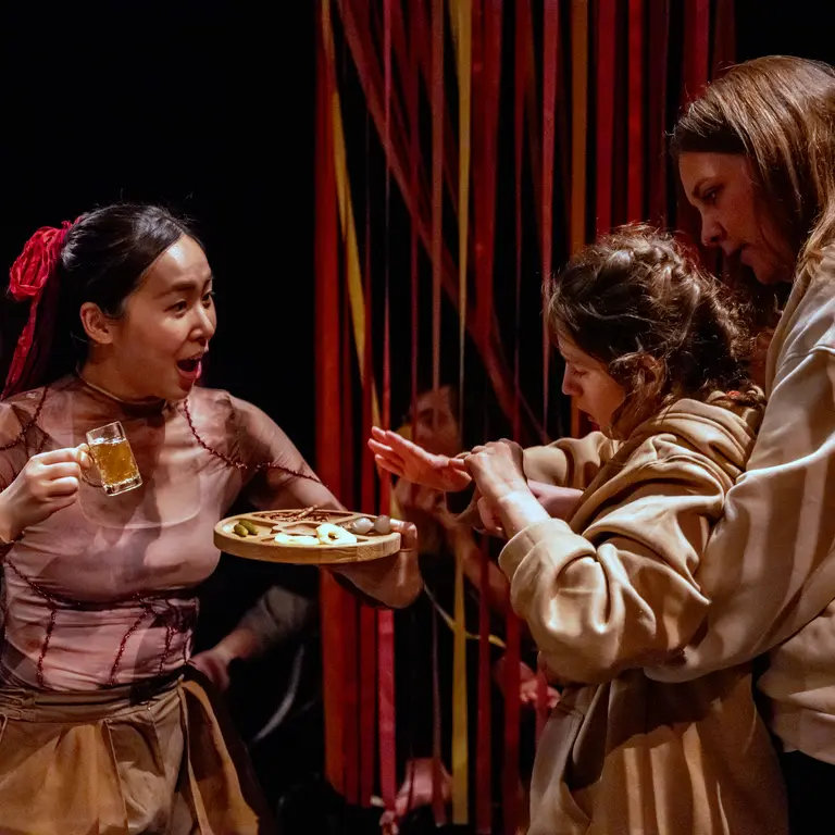 Three performers interact on stage, with one holding a tray and mug while two others lean in closely, surrounded by hanging ribbons and theatrical lighting during a live performance scene.