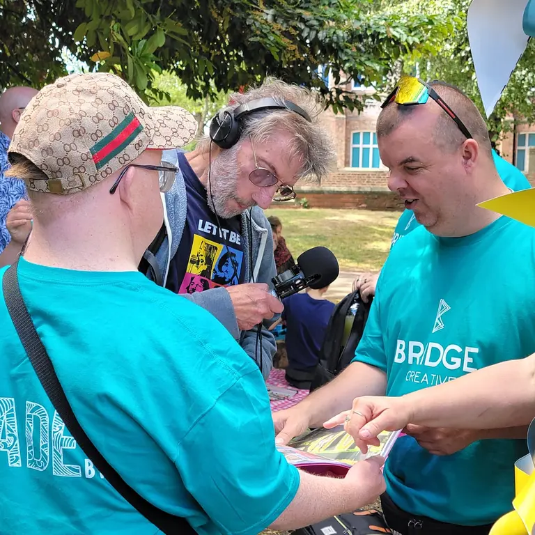 Participants at a Bridge Creative outdoor session gather around a table, exchanging printed materials while recording audio, with colourful crafted decorations hanging nearby.