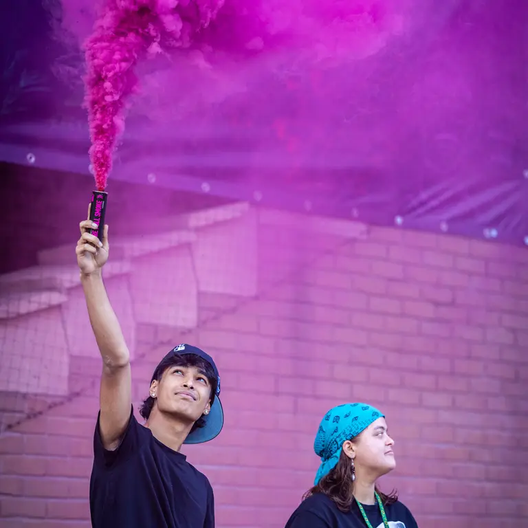 two young people with blue backward caps and dark blue shirts let off a purple flare