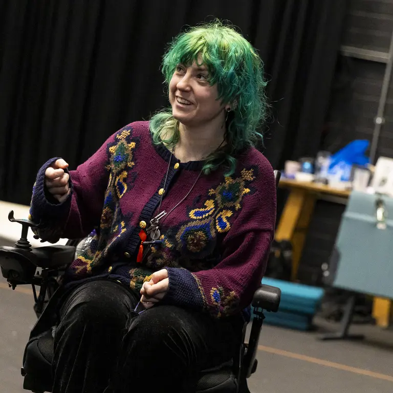 A participant using a powered wheelchair takes part in a rehearsal space, gesturing with one hand while seated on stage, with props and equipment visible in the background.