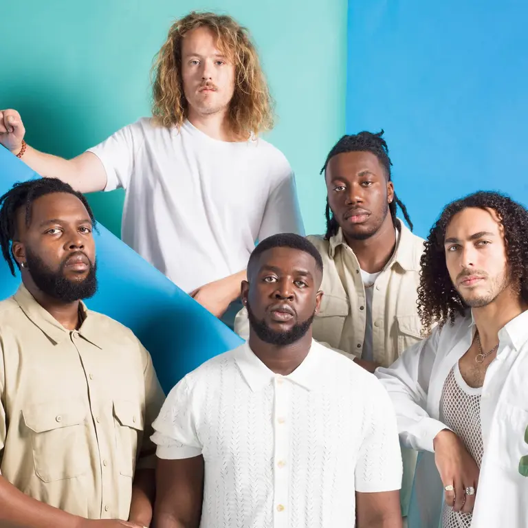 Ezra Collective band members wearing white/cream tops on a blue staircase and staring at the camera with no expression