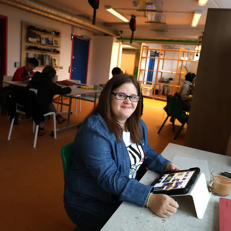A woman sits in a public space with a tablet and smiles towards the camera