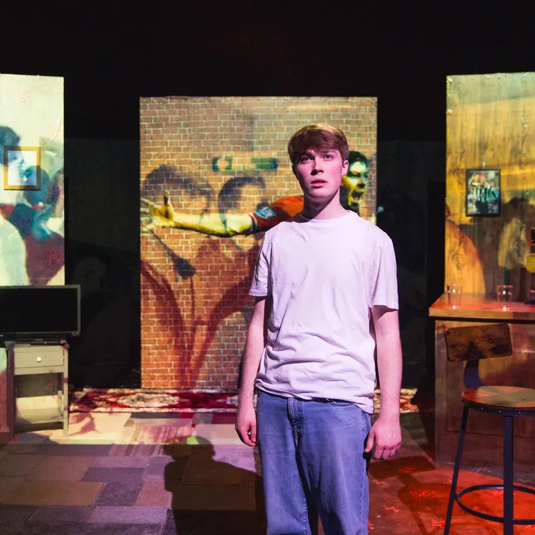 A young man stands on stage looking into the distance, while images are projected onto screens behind him