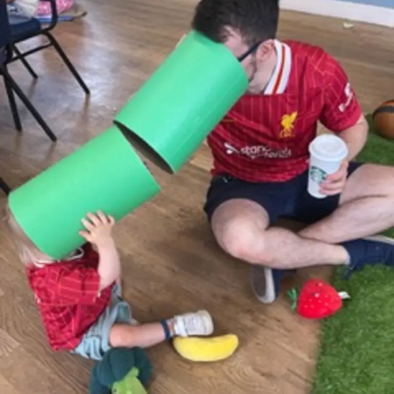 A young child and a man at on the floor hold up large green tubes to their faces, they peer down the tubes at each other. 